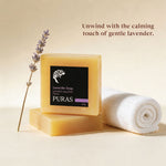 A bar of PURAS lavender soap stands on another bar, with a rolled white towel and a lavender sprig beside it on a warm beige background. Text reads: 'Unwind with the calming touch of gentle lavender.