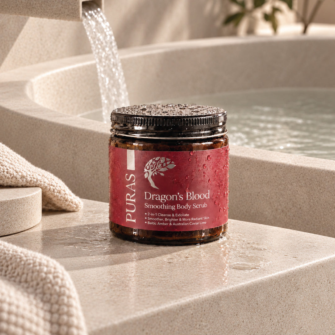 PURAS Dragon’s Blood Smoothing Body Scrub in an amber jar with a maroon label, on a wet stone ledge beside a bath.