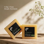 Two bars of PURAS Lemongrass Soap displayed on a wooden surface with a warm beige background and soft shadows. One bar is standing upright and the other is leaning on its side, both showing their black labels.