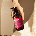 A hand holding a dark amber pump bottle of PURA'S Dragon's Blood Body Lotion against a warm beige background with a textured cream towel.
