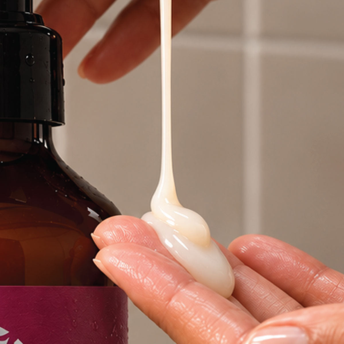 Close-up of creamy white lotion being dispensed onto fingertips beside an amber pump bottle with a maroon label.
